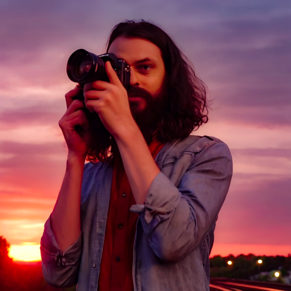 Rito in a sunset background holding a camera to his eye looking through the lens