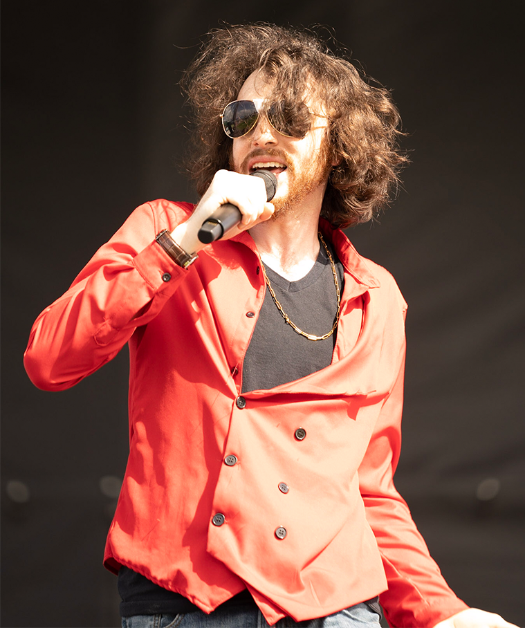 Rito Rhymes holding a microphone to his mouth rapping wearing a red shirt and sunglasses with tape in the middle