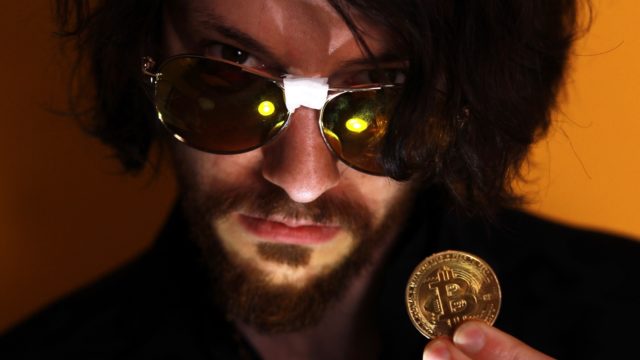 Rito in a close up holding a Bitcoin coin and looking into the camera with sunglasses with tape in the middle; shadowy image